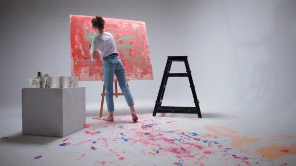 Νεαρή γυναίκα καλλιτέχνης ζωγραφίζει με ένα πινέλο σε ένα μεγάλο καμβά σε ένα λευκό δωμάτιο, ένας ταλαντούχος καλλιτέχνης αντλεί ένα χρώμα αφαίρεσης, σύγχρονη τέχνη στη ζωγραφική πετρελαίου. — Αρχείο Βίντεο