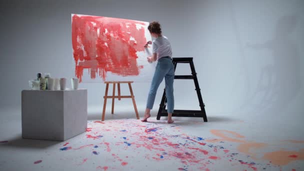 Visão traseira, artista mulher desenha com um pincel em uma tela grande em uma sala branca, um artista talentoso desenha uma abstração de cor vermelha, arte moderna em pintura a óleo. — Vídeo de Stock