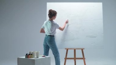 Arkaya bakan kadın sanatçı beyaz bir odadaki büyük bir tuvale beyaz bir astar uyguluyor, yetenekli bir sanatçı çizmeye hazırlanıyor, 4k yavaş çekim..