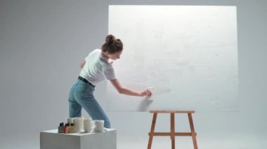 Kadın sanatçı beyaz bir odadaki büyük bir tuvale beyaz astar katmanı uyguluyor, yetenekli bir sanatçı çizmeye hazırlanıyor, 4k yavaş çekim..