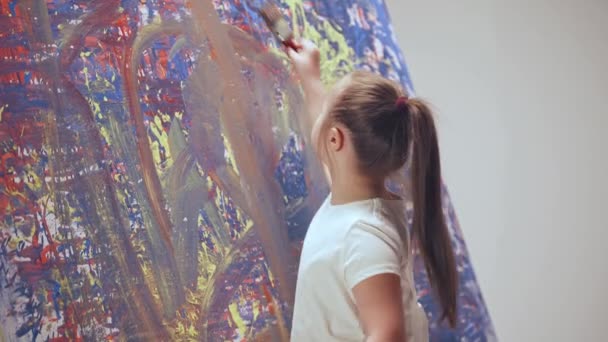 Девушка с особыми потребностями рисует кистью на большом холсте в белой комнате, девочка с синдромом Дауна рисует красно-голубую абстракцию, вид сзади. — стоковое видео
