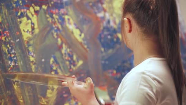 Fille avec des besoins spéciaux dessine avec un pinceau sur une grande toile dans une chambre blanche, enfant fille avec le syndrome du duvet dessine une abstraction couleur rouge-bleu, vue de dos. — Video