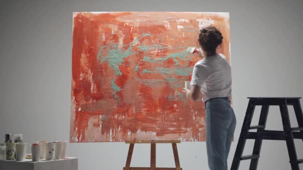 Γυναίκα καλλιτέχνης ζωγραφίζει με ένα πινέλο σε ένα μεγάλο καμβά σε ένα λευκό δωμάτιο, ένας ταλαντούχος καλλιτέχνης σχεδιάζει ένα κόκκινο χρώμα αφαίρεσης, πιτσιλίζοντας χρώμα σε καμβά. — Αρχείο Βίντεο