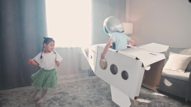 Chinese kids play in the living room of the house, a boy in an astronaut costume plays with a cardboard space shuttle, a cute girl sister runs after her brother around the room, 4k slow motion. — Vídeos de Stock