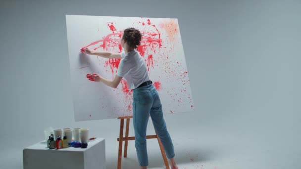 Γυναίκα καλλιτέχνης ζωγραφίζει με τα χέρια της σε ένα μεγάλο καμβά σε ένα λευκό δωμάτιο, ένας ταλαντούχος καλλιτέχνης ζωγραφίζει ένα κόκκινο χρώμα αφαίρεσης, αντλεί με πιτσιλίσματα χρωμάτων. — Αρχείο Βίντεο