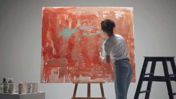 女性アーティストは白い部屋の大きなキャンバスにブラシで描く、才能のあるアーティストは、色の赤の抽象化を描き、キャンバスにペイントをはね、バックビュー. — ストック動画