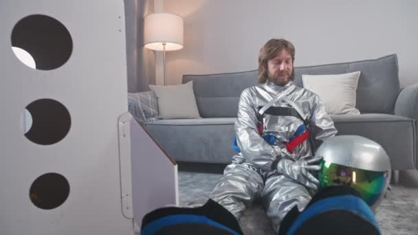 Man in an astronaut suit sits on the floor in living room next to a cardboard model of a space shuttle, an astronaut puts on a helmet, a childs dream. — Stok video