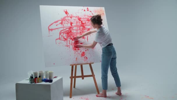 Jeune femme artiste dessine avec ses mains sur une grande toile dans une pièce blanche, une artiste talentueuse dessine une abstraction de couleur rouge, dessine par éclaboussures de peinture. — Video