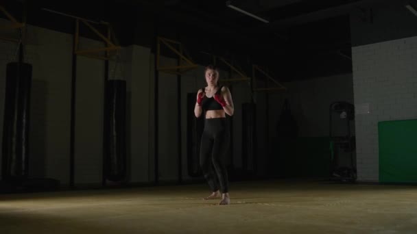 Kobieta bokser trenuje swoje kopnięcia, trening w siłowni bokserskiej, młoda kobieta patrzy w kamerę i trenuje serię ciosów. — Wideo stockowe