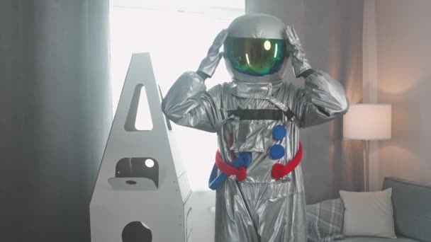 Kosmonauta, mężczyzna w średnim wieku w stroju astronauty stoi w salonie obok tekturowego modelu wahadłowca, astronauta odkłada hełm, dziecięcy sen, 4k zwolniony ruch. — Wideo stockowe