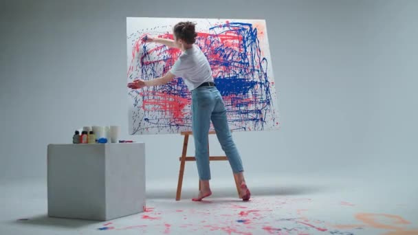 女性アーティストは白い部屋の大きなキャンバスに絵の具を撒き散らし、才能あるアーティストは赤青の抽象画を描きます。. — ストック動画