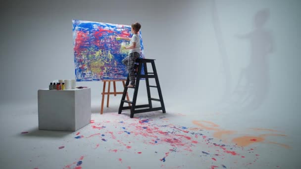 Malý chlapec kreslí štětcem na velké plátno v bílém pokoji, dětský umělec kreslí barevnou abstrakci, stříkající malbu na plátno. — Stock video