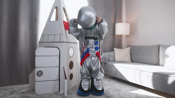 Azjata w kostiumie astronauty stoi w pobliżu kartonowego modelu statku kosmicznego i zakłada hełm, patrzy w kamerę i fale, chłopiec gra astronautę w salonie domu. — Wideo stockowe