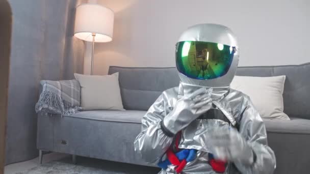 Man in an astronaut suit sits on the floor in living room next to a cardboard model of a space shuttle, an astronaut opens the visor in his helmet and looks at the shuttle, a childs dream. — Vídeo de Stock