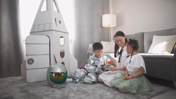 Femme asiatique avec des enfants jouent dans le salon à la maison, un garçon en costume d'astronaute assis sur le sol avec sa mère et sa sœur, les enfants jouent avec un modèle de jouet du système solaire, 4k au ralenti — Video