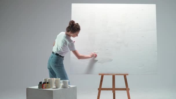 Vista posterior, artista femenina aplica una capa de imprimación blanca sobre un lienzo grande en una habitación blanca, un artista talentoso que se prepara para dibujar. — Vídeo de stock