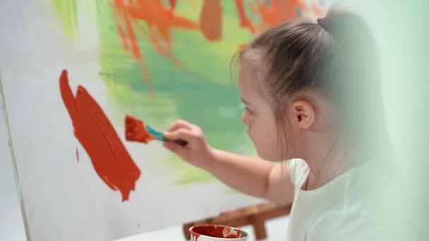 Menina com síndrome de down desenha com uma escova em uma tela grande em uma sala branca, menina criança com necessidades especiais desenha uma abstração de cor, 4k câmera lenta. — Vídeo de Stock