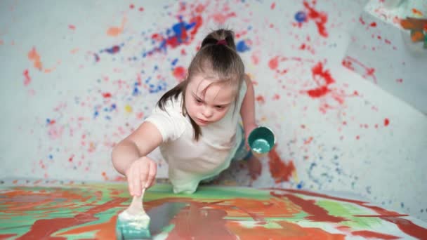 Meisje met down syndroom tekent met een borstel op een groot doek in een witte kamer, meisje met speciale behoeften tekent een kleur abstractie, bovenaanzicht, 4k slow motion. — Stockvideo