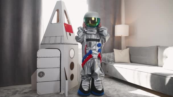 Un chico asiático disfrazado de astronauta se para cerca de un modelo de cartón de una nave espacial y se quita el casco, mira a la cámara y las olas, un niño interpreta a un astronauta en la sala de estar de la casa. — Vídeo de stock