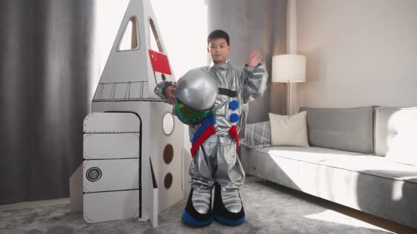 Un chico asiático disfrazado de astronauta se para cerca de un modelo de cartón de una nave espacial y se pone el casco, mira a la cámara y las olas, un niño interpreta a un astronauta en la sala de estar de la casa. — Vídeo de stock