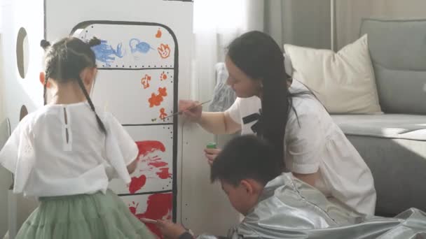 Una mujer asiática con niños jugando en la sala de estar en casa, un niño con un disfraz de astronauta sentado en el suelo con su madre y su hermana, niños junto con su madre pintan en un modelo de cartón — Vídeo de stock