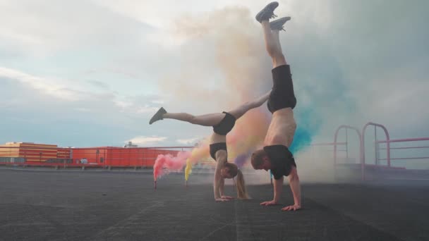 Slow motion, kvinna och man gör en handstand, mannen gör en volt, flerfärgade rökbomber på en bakgrund, gymnastiska element och parkour på taket av byggnaden. — Stockvideo