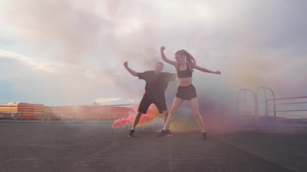 Slow motion, kvinna och man gör en kullerbytta, flerfärgade rökbomber på en bakgrund, gymnastiska element och parkour på taket av byggnaden. — Stockvideo