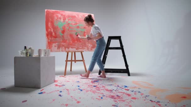Joven artista femenina dibuja con un pincel en un gran lienzo en una habitación blanca, un artista talentoso dibuja una abstracción de color, arte moderno en la pintura al óleo. — Vídeo de stock