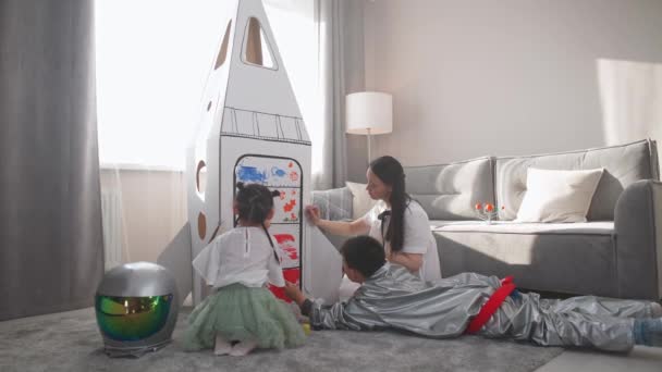 Çocuklu Asyalı bir kadın evdeki oturma odasında oynuyor. Astronot kostümlü bir çocuk yerde annesi ve kız kardeşiyle oturuyor. Çocuklar anneleriyle birlikte karton bir modelin üzerinde. — Stok video