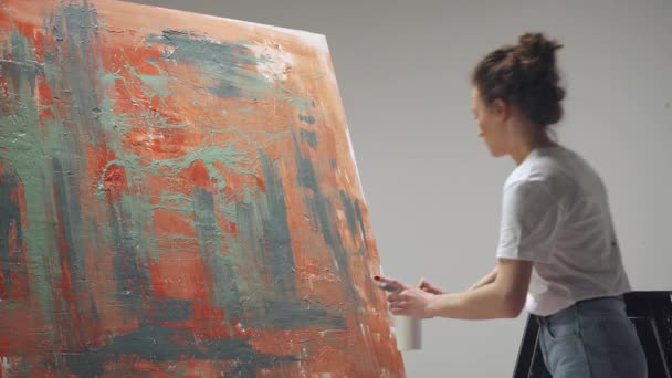 Jeune femme artiste dessine avec un pinceau sur une grande toile dans une pièce blanche, un artiste talentueux dessine une abstraction de couleur rouge, éclaboussure de peinture sur toile, vue de dos. — Video