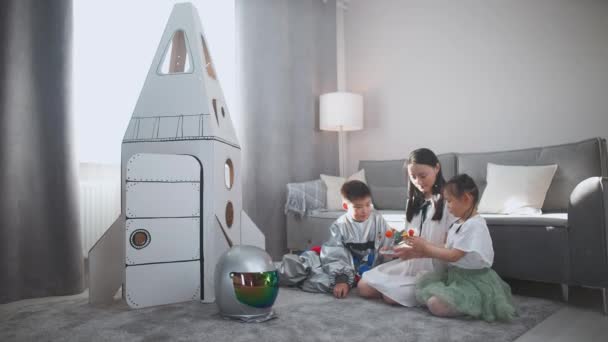 Asian mother with kids play in the living room at home, a boy in an astronaut costume sitting on the floor with her mother and sister, woman tells her children about the solar system, study of — Vídeo de Stock