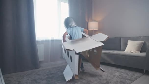 Мальчик в костюме космонавта играет с картонным космическим шаттлом, бегает по комнате, 4k замедленной съемки. — стоковое видео