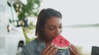 Doğada neşeli bir Afrikalı kadın portresi, karpuz yiyen ve meyve yemeğinin tadını çıkaran genç bir kadın, yaz, doğada piknik, 4k yavaş çekim.