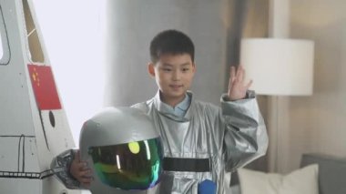 Astronot kostümü giymiş Asyalı bir çocuk uzay gemisinin karton modelinin yanında duruyor ve kaskını takıyor, kameraya bakıyor ve dalgalara bakıyor, bir çocuk evin oturma odasında astronotu oynuyor..