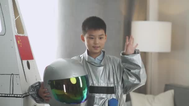 Un chico asiático disfrazado de astronauta se para cerca de un modelo de cartón de una nave espacial y se pone el casco, mira a la cámara y las olas, un niño interpreta a un astronauta en la sala de estar de la casa. — Vídeos de Stock