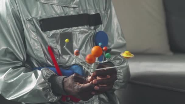 African man in an astronaut costume sitting on the floor in living room at home and playing with a toy model of the solar system, study of astronomy, man on self-isolation. — Stock Video