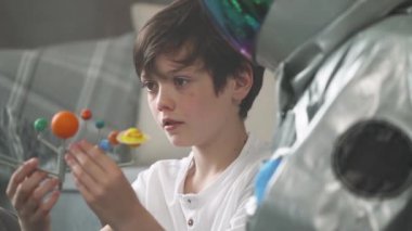 Küçük çocuk evdeki oturma odasında oynuyor, astronot kostümlü bir adam oğluyla yerde oturuyor, güneş sisteminin oyuncak modeliyle oynuyor, astronomi okuyor..