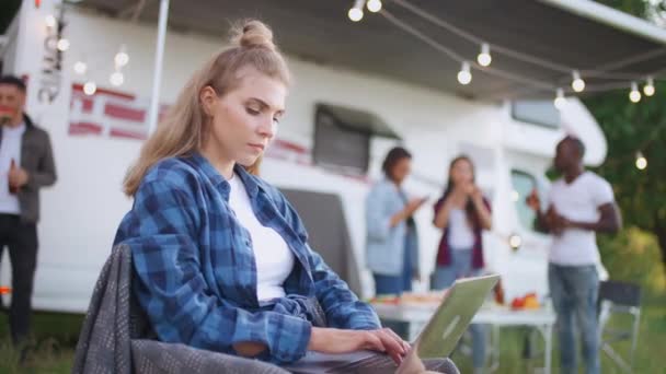 Caucasico freelance femminile che lavora presso un computer portatile mentre seduto vicino camper, digitando testo e lavoro a distanza nella natura, donna concentrata lavora mentre in vacanza, amici e giovani si rilassano nella — Video Stock