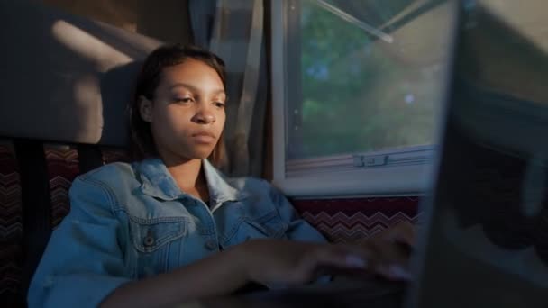 Afrykanka freelancer pracuje na laptopie podczas siedzenia w samochodzie, wpisując tekst i zdalnej pracy w przyrodzie, portret skupionej kobiety w świetle zachodu słońca wewnątrz samochodu kempingowego. — Wideo stockowe
