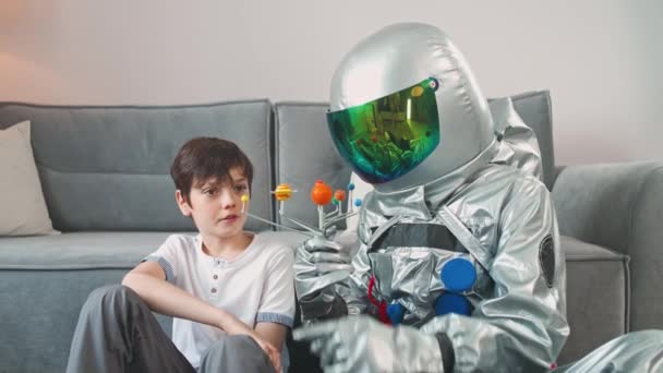 Ojciec i syn bawią się w salonie w domu, ojciec w kostiumie astronauty siedzący na podłodze z jej synem, chłopiec bawiący się zabawkowym modelem Układu Słonecznego, nauka astronomii, 4k slow motion. — Wideo stockowe