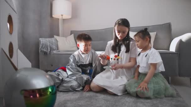 Asiatisk mamma med barn leker i vardagsrummet hemma, en pojke i en astronaut kostym sitter på golvet med sin mor och syster, kvinna berättar för sina barn om solsystemet. — Stockvideo