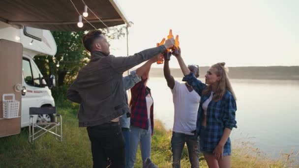 L'été, un groupe de jeunes profitent d'une fête dans la nature près du lac, les jeunes de différentes races boivent de la bière et se relaxent dans la nature près du camping-car, coucher de soleil. — Video