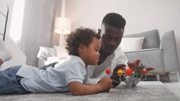 Afrikaanse familie, vader en zoon spelen in de woonkamer thuis, man ligt op de vloer met haar zoon, jongen speelt met een speelgoedmodel van het zonnestelsel, studie van astronomie, 4k slow motion. — Stockvideo