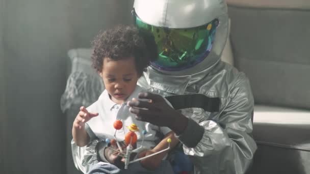 Afrikai apa és fia játszanak a nappaliban otthon, férfi űrhajós jelmezben ül a földön a fiával, fiú játszik egy játék modell a naprendszer, csillagászat. — Stock videók