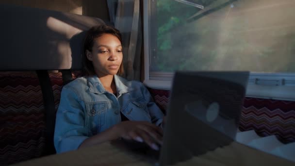 Femme indépendante africaine travaillant à un ordinateur portable assis dans une voiture, tapant du texte et du travail à distance dans la nature, portrait d'une femme concentrée dans une lumière du coucher du soleil à l'intérieur d'un camping-car, 4k au ralenti. — Video