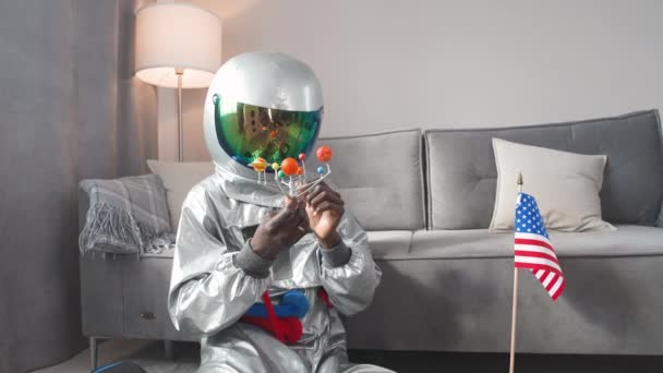 Afrikansk man i en astronaut kostym sitter på golvet i vardagsrummet hemma och leker med en leksak modell av solsystemet, studier av astronomi, man på självisolering, 4k slow motion. — Stockvideo