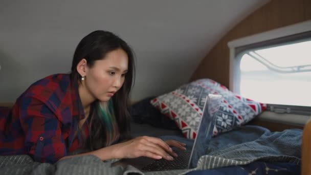 Femme asiatique pigiste travaillant sur un ordinateur portable alors qu'elle était allongée dans une maison mobile, tapant du texte, travaillant à distance dans la nature, portrait à l'intérieur d'une voiture de camping. — Video