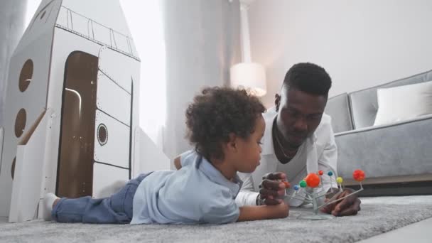 Ayah dan anak Afrika bermain di ruang tamu di rumah, laki-laki tergeletak di lantai dengan anaknya, anak laki-laki bermain dengan mainan model tata surya, studi astronomi, gerakan lambat 4k. — Stok Video