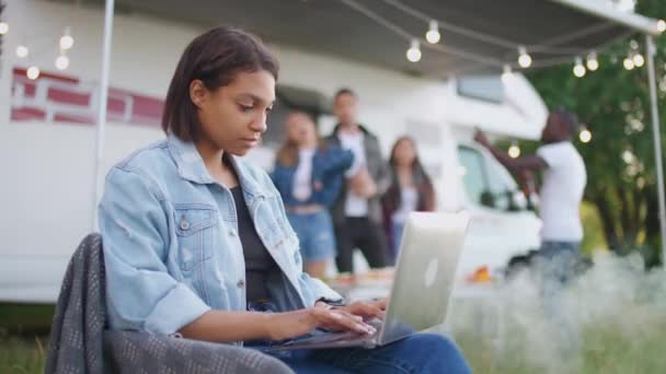 Female freelance working at a laptop while sitting near camping car, typing text and remote work in nature, friends and young people relax in the background, a party, african female pauses work and — Stock Video