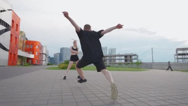 Young man doing a somersaults on training area near modern buildings, guy and a girl perform gymnastic tricks, 4k slow motion. — Stock Video